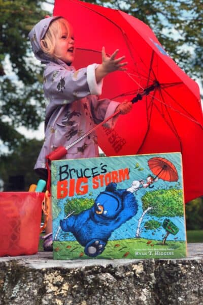 little girl in purple rain jacket holding red umbrella standing on a tree stump with a bucket and a book called "bruce's Big storm" in the foreground